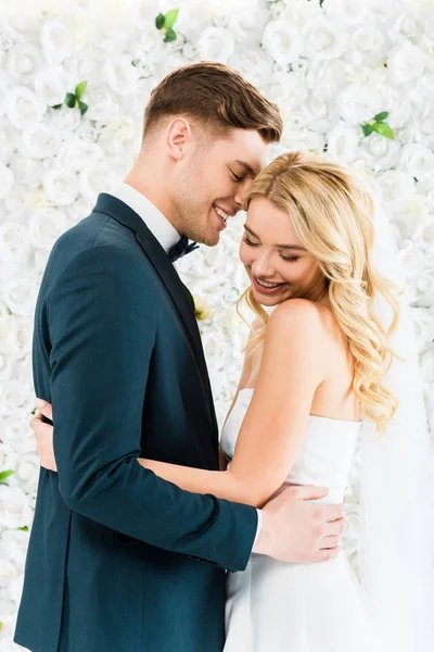 Feliz hermosa novia abrazando novio sonriente sobre fondo floral blanco - foto de stock