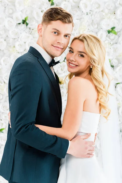 Усміхнена красива наречена обіймає гарного нареченого на білому квітковому тлі — стокове фото