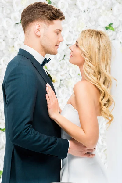 Счастливая молодая невеста и жених стоя лицом к лицу на белом цветочном фоне — стоковое фото