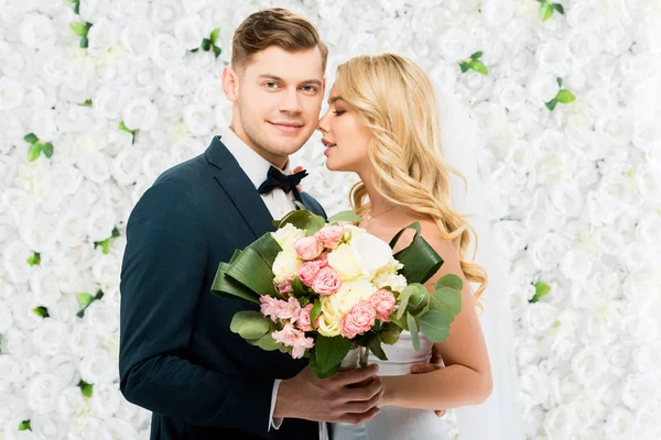 Heureux jeune mariée et marié tenant bouquet de mariage sur fond floral blanc — Photo de stock