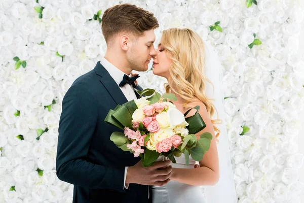 Felice giovane sposa e sposo baciare mentre tiene bouquet da sposa su sfondo floreale bianco — Foto stock