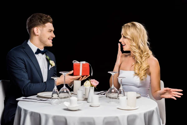 Happy handsome groom presenting red gift box to surprised bride, while sitting at served table isolated on black — Stock Photo