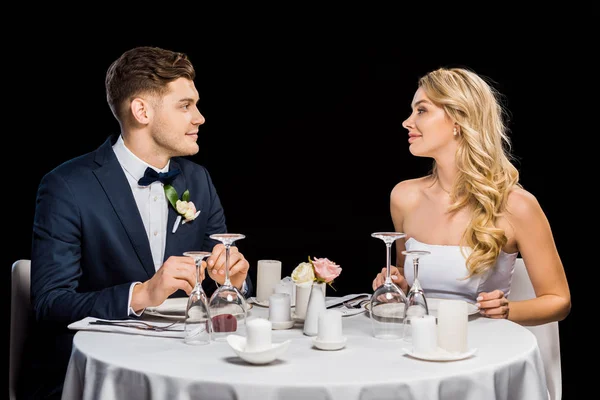 Handsome groom and beautiful bride sitting at served table isolated on black — Stock Photo