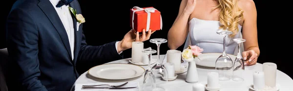 Plan panoramique du marié présentant boîte cadeau rouge à la mariée, tout en étant assis à la table servie isolé sur noir — Photo de stock