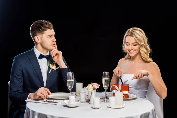 Novio desanimado mirando a la novia con caja de regalo en el plato aislado en negro - foto de stock