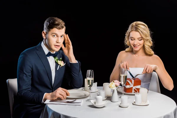 Novio desanimado mirando a la cámara mientras la novia con caja de regalo en el plato imitando comer aislado en negro - foto de stock