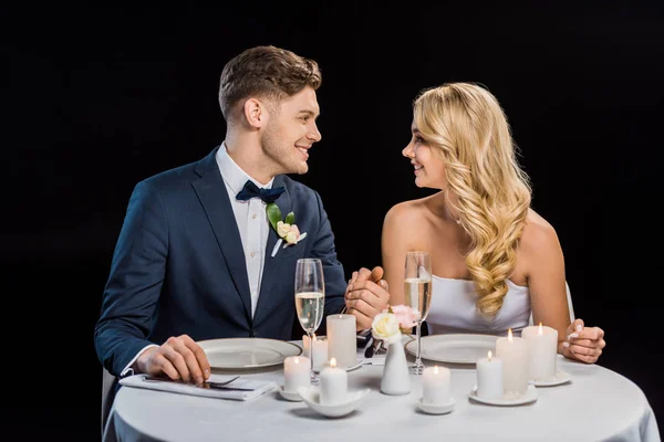 Glücklicher junger Bräutigam und Braut Händchen haltend, während sie am gedeckten Tisch isoliert auf schwarz sitzen — Stockfoto