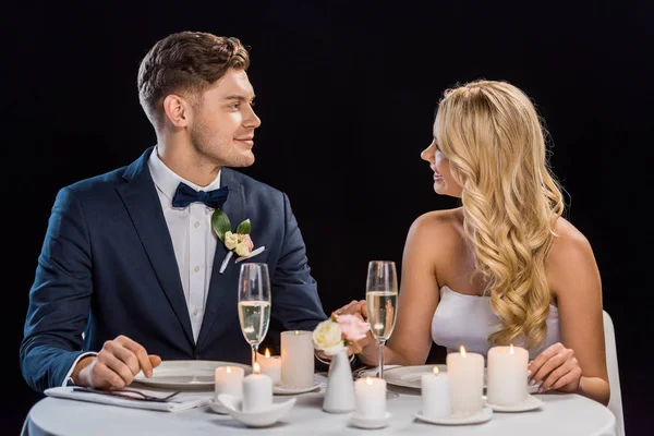 Feliz joven pareja sentado a la mesa servida y mirándose el uno al otro aislado en negro - foto de stock