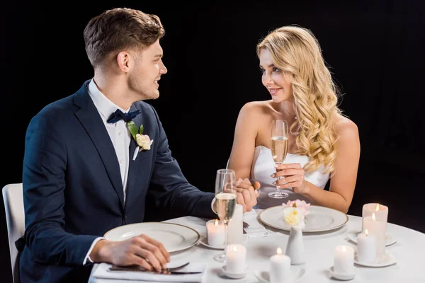 Feliz joven pareja sentada en la mesa servida con copas de champán y mirándose aislados en negro - foto de stock