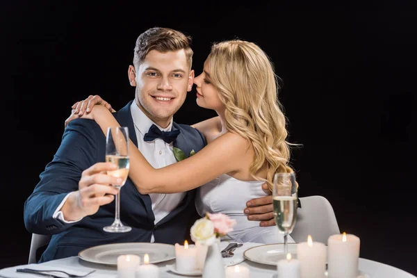 Heureuse jeune mariée embrassant beau marié avec verre de champagne isolé sur noir — Photo de stock