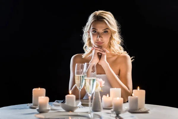 Beautiful young bride sitting alone at table with burning candles isolated on black — Stock Photo