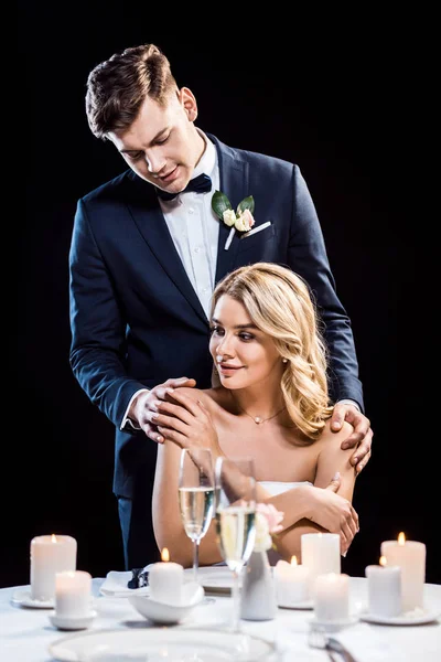 Smiling groom hugging pretty bride from back by shoulders isolated on black — Stock Photo