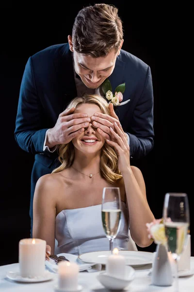 Smiling groom standing behind sitting bride and closing her eyes with hands isolated on black — Stock Photo