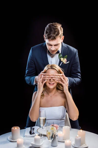 Young groom standing behind sitting bride and closing her eyes with hands isolated on black — Stock Photo