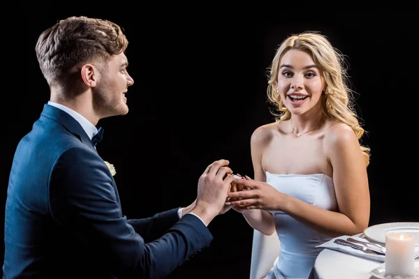 Feliz joven hombre haciendo matrimonio propuesta a alegre joven mujer aislado en negro - foto de stock