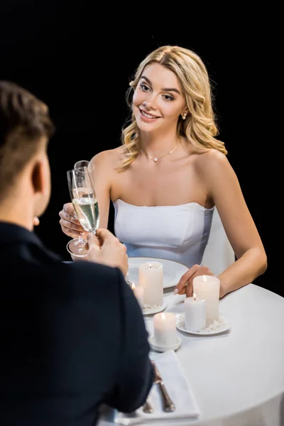 Enfoque selectivo de pareja joven tintineando copas de champán mientras está sentado en la mesa servida aislado en negro - foto de stock