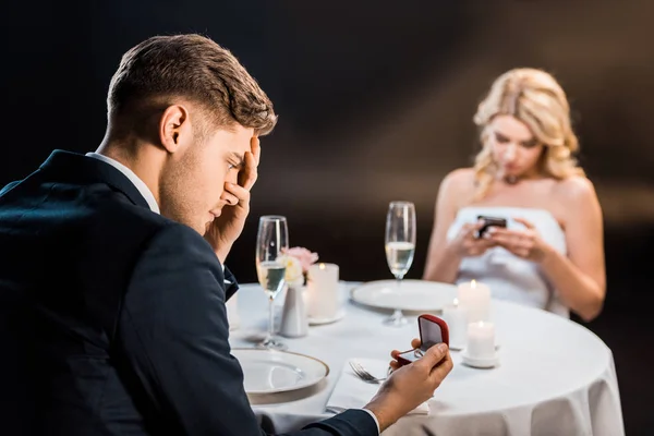 Selective focus of sad young man holding gift box with wedding ring while girl using smartphone on black background — Stock Photo