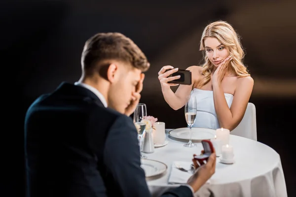 Messa a fuoco selettiva di bella giovane donna scattare selfie con smartphone, mentre l'uomo triste in possesso di scatola regalo con fede nuziale su sfondo nero — Foto stock