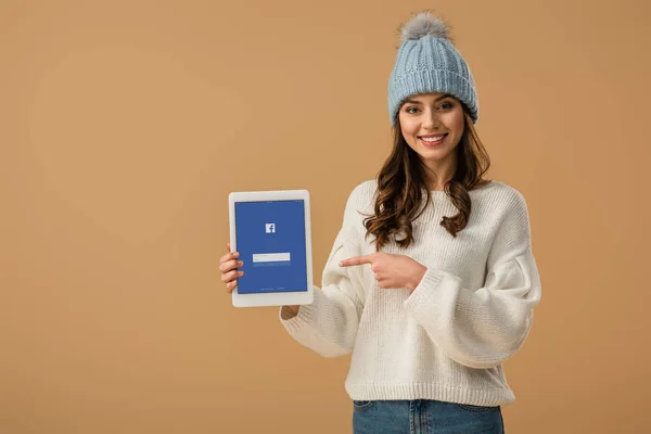 Wunderschöne brünette Mädchen mit Strickmütze zeigt mit dem Finger auf digitales Tablet mit Facebook-App auf dem Bildschirm isoliert auf beige — Stockfoto