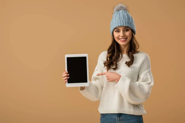 Lachende, lockige Frau mit Strickmütze, die mit dem Finger auf ein digitales Tablet mit leerem Bildschirm zeigt, isoliert auf beige — Stockfoto