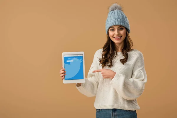 Mujer morena sonriente en suéter blanco apuntando con el dedo a la tableta digital con aplicación de twitter en pantalla aislada en beige - foto de stock
