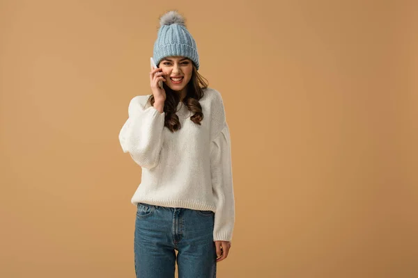 Chica morena divertida en sombrero de punto hablando en teléfono inteligente aislado en beige - foto de stock