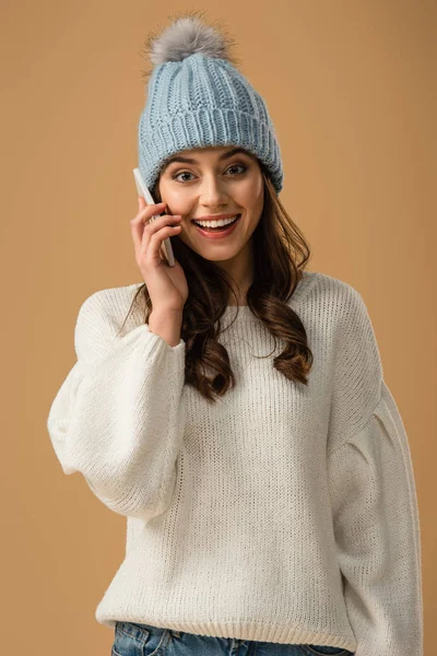 Брюнетка в вязаной шляпе разговаривает на смартфоне с удивленной улыбкой, изолированной от бежевого — стоковое фото