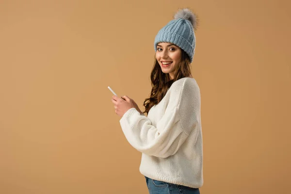 Excited curly woman in knitted hat holding smartphone isolated on beige — Stock Photo