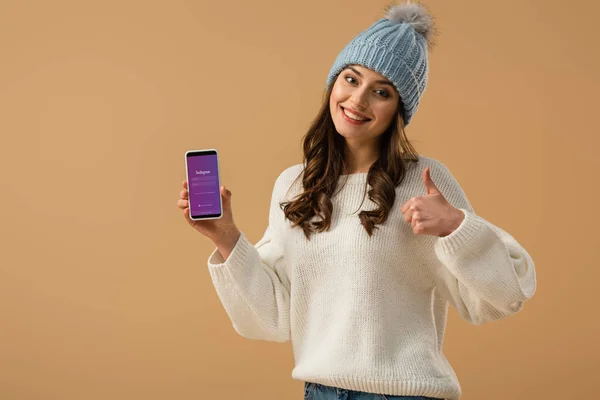 Fröhlich lächelndes Mädchen im Pullover hält Smartphone mit Instagram-App auf dem Bildschirm und zeigt Daumen hoch vereinzelt auf beige — Stockfoto