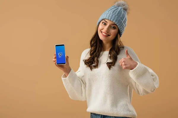 Chica morena feliz sosteniendo teléfono inteligente con aplicación shazam en la pantalla y mostrando el pulgar hacia arriba aislado en beige - foto de stock