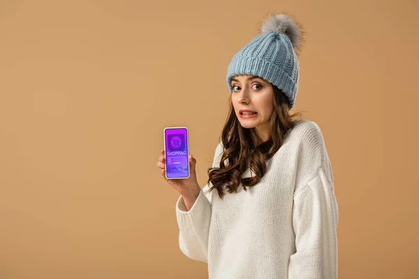 Fille brune confus dans le chapeau tenant smartphone avec application shopping à l'écran isolé sur beige — Photo de stock