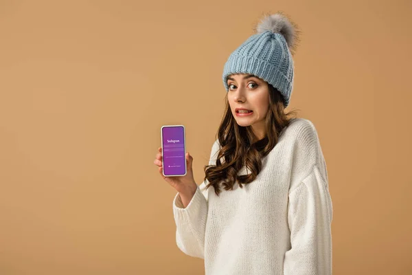 Chica atractiva confusa sosteniendo teléfono inteligente con aplicación instagram en la pantalla aislada en beige - foto de stock