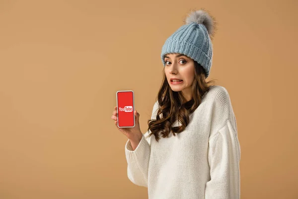 Chica bastante confundida sosteniendo teléfono inteligente con aplicación de youtube en la pantalla aislada en beige - foto de stock