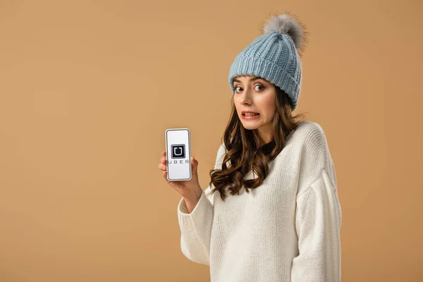 Confused brunette girl in sweater holding smartphone with uber app on screen isolated on beige — Stock Photo