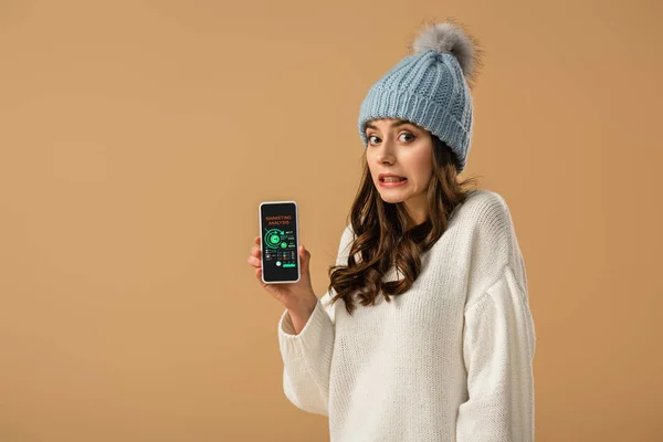 Pretty confused girl in sweater holding smartphone with marketing analysis app on screen isolated on beige — Stock Photo