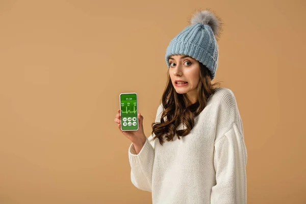 Confused curly young woman holding smartphone with health app on screen isolated on beige — Stock Photo