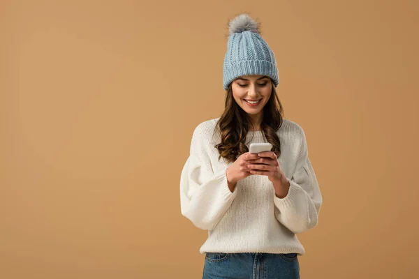 Cheerful brunette woman in white sweater using smartphone isolated on beige — Stock Photo
