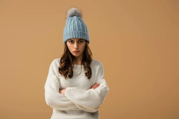 Mujer morena trastornada en sombrero de punto de pie con brazos cruzados aislados en beige - foto de stock