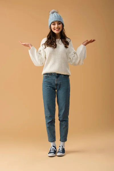 Vista completa de la chica morena riendo en jeans y suéter de pie sobre fondo beige - foto de stock