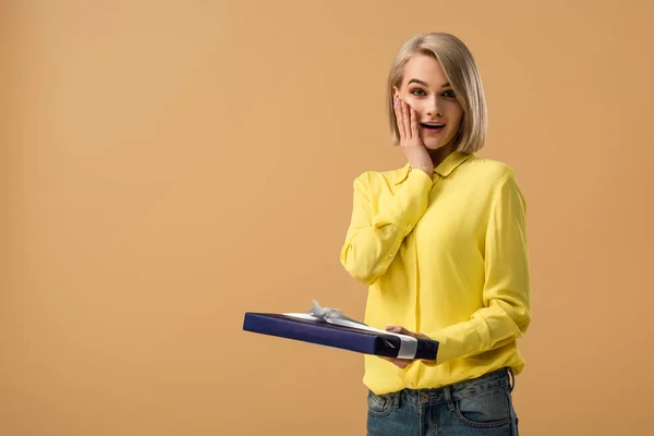 Surprised blonde woman in yellow shirt touching face and holding gift box isolated on beige — Stock Photo