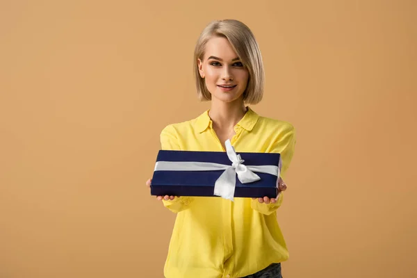 Pretty blonde young woman showing gift box with ribbon isolated on beige — Stock Photo