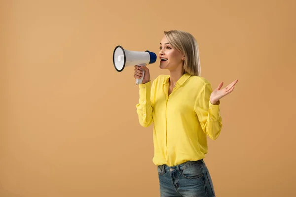 Femme blonde souriante en chemise jaune criant en mégaphone isolé sur beige — Photo de stock