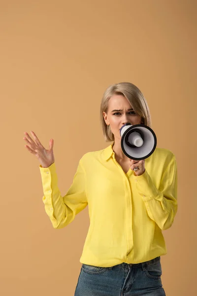 Worried blonde woman in yellow shirt talking in megaphone isolated on beige — Stock Photo