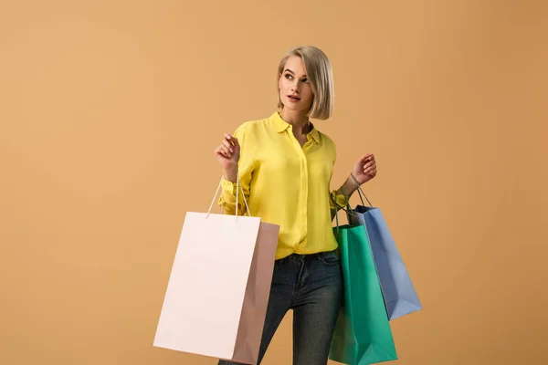 Pensive blonde girl in yellow shirt holding shopping bags isolated on beige — Stock Photo