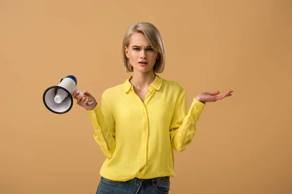 Confused blonde girl in yellow shirt holding loudspeaker isolated on beige — Stock Photo