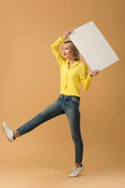 Ganzkörperansicht der blonden Frau in Jeans, die auf einem Bein steht und ein leeres Plakat auf beigem Hintergrund hält — Stockfoto