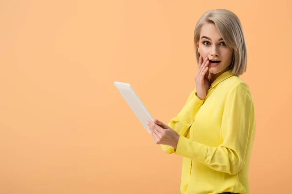 Shocked blonde woman in yellow shirt holding digital tablet isolated on orange — Stock Photo