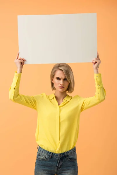 Dissatisfied blonde girl in yellow shirt holding blank placard isolated on orange — Stock Photo