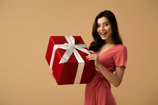 Chica morena sorprendida en vestido sosteniendo caja de regalo roja y sonriendo aislado en beige - foto de stock