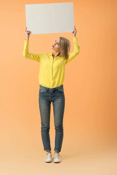 Full length view of irritated blonde girl in jeans holding blank placard on orange background — Stock Photo